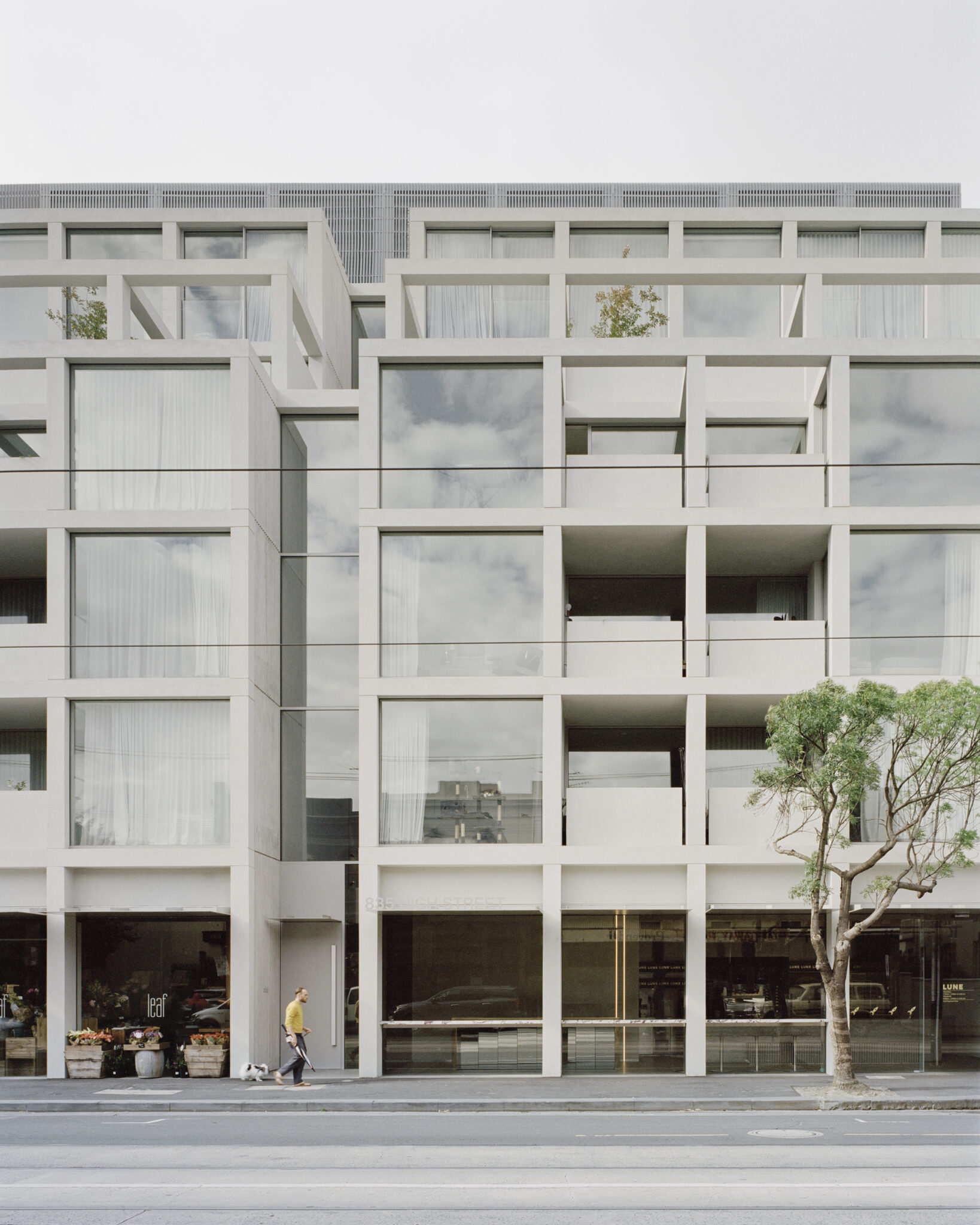 Modern multi-storey mixed-use building with a white geometric façade, large windows and balconies, ground-floor retail, street trees and pedestrians along a tram-lined urban street.