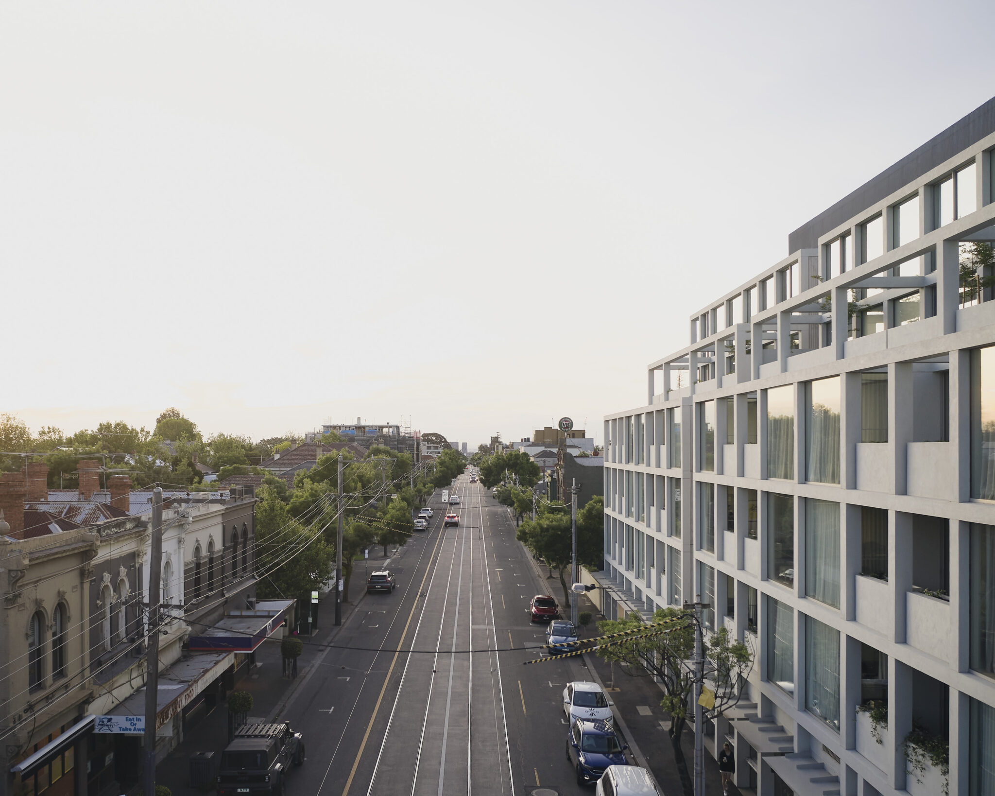 Modern multi-storey building overlooking a tram-lined urban street, with a mix of historic low-rise buildings, established trees and residential development extending into the distance.