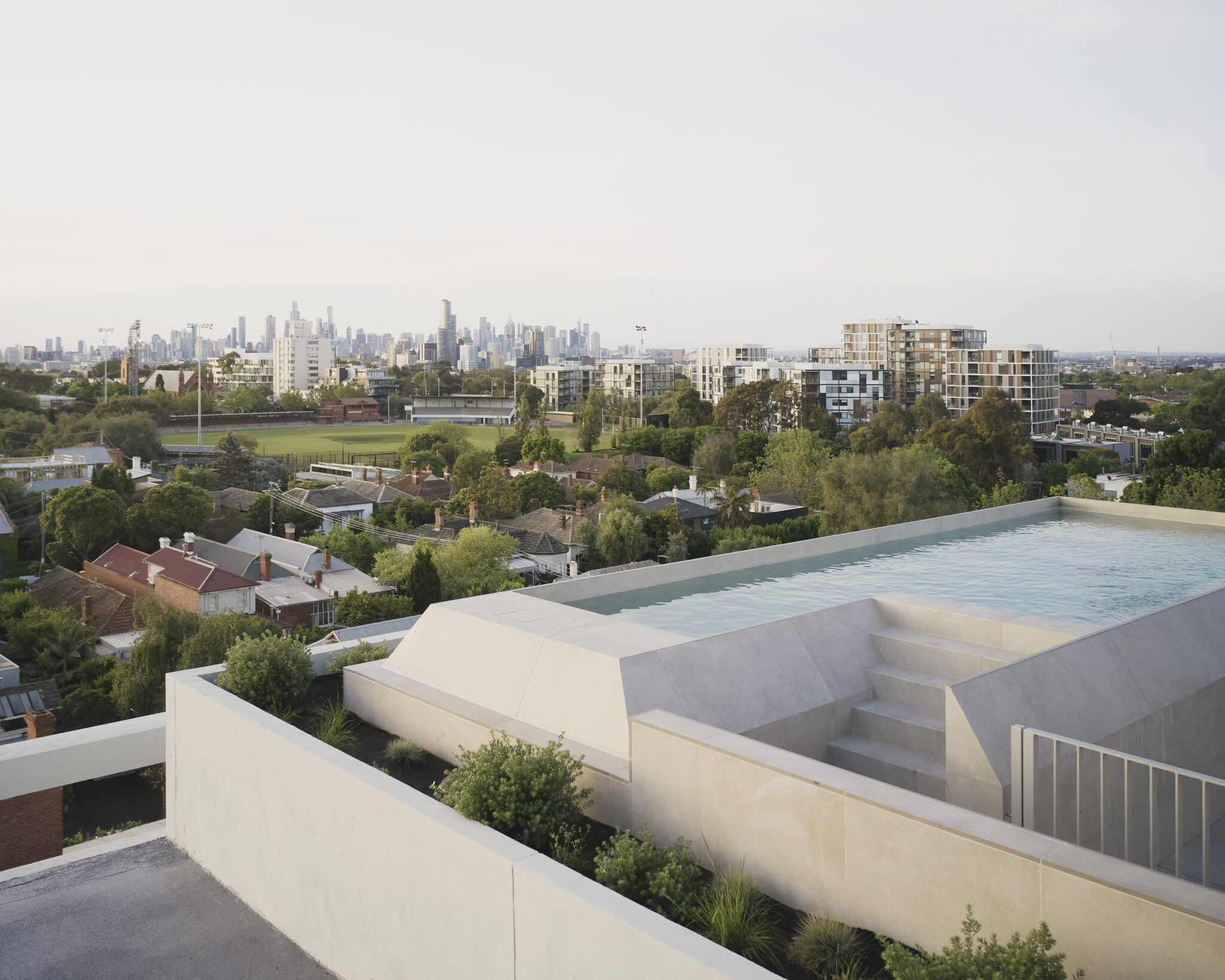 Modern rooftop terrace with an infinity pool overlooking a suburban Melbourne landscape, with established housing, green open space and a dense city skyline in the distance.