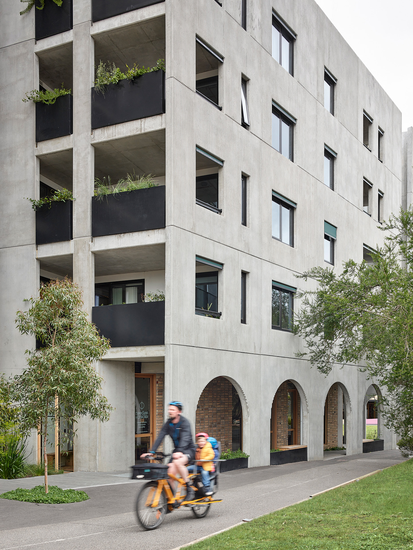 Modern apartment building with concrete façade, planted balconies and arched ground-floor openings, with a cyclist and child riding along a landscaped pedestrian path.