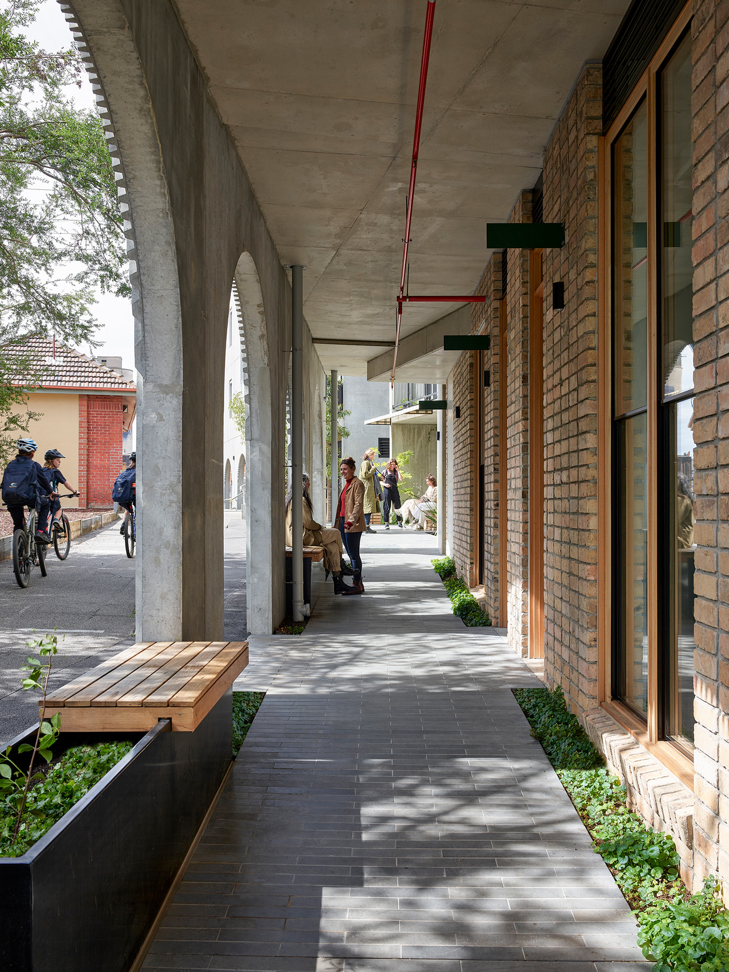 Covered pedestrian arcade with concrete arches and brick storefronts, people sitting and chatting on benches while cyclists pass by, illustrating a sheltered, active neighbourhood streetscape.
