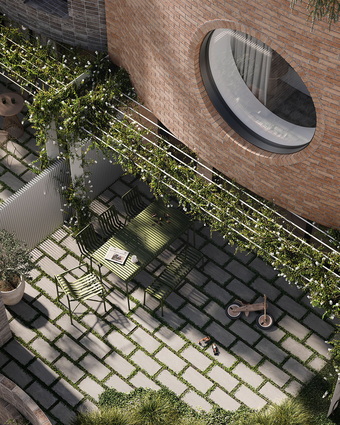 Sunlit private courtyard for dining and play with a round window set in a brick wall, a green metal table and chairs beneath a vine-covered pergola, concrete pavers with grass joints, and a wooden balance bike and small toy on the patio.