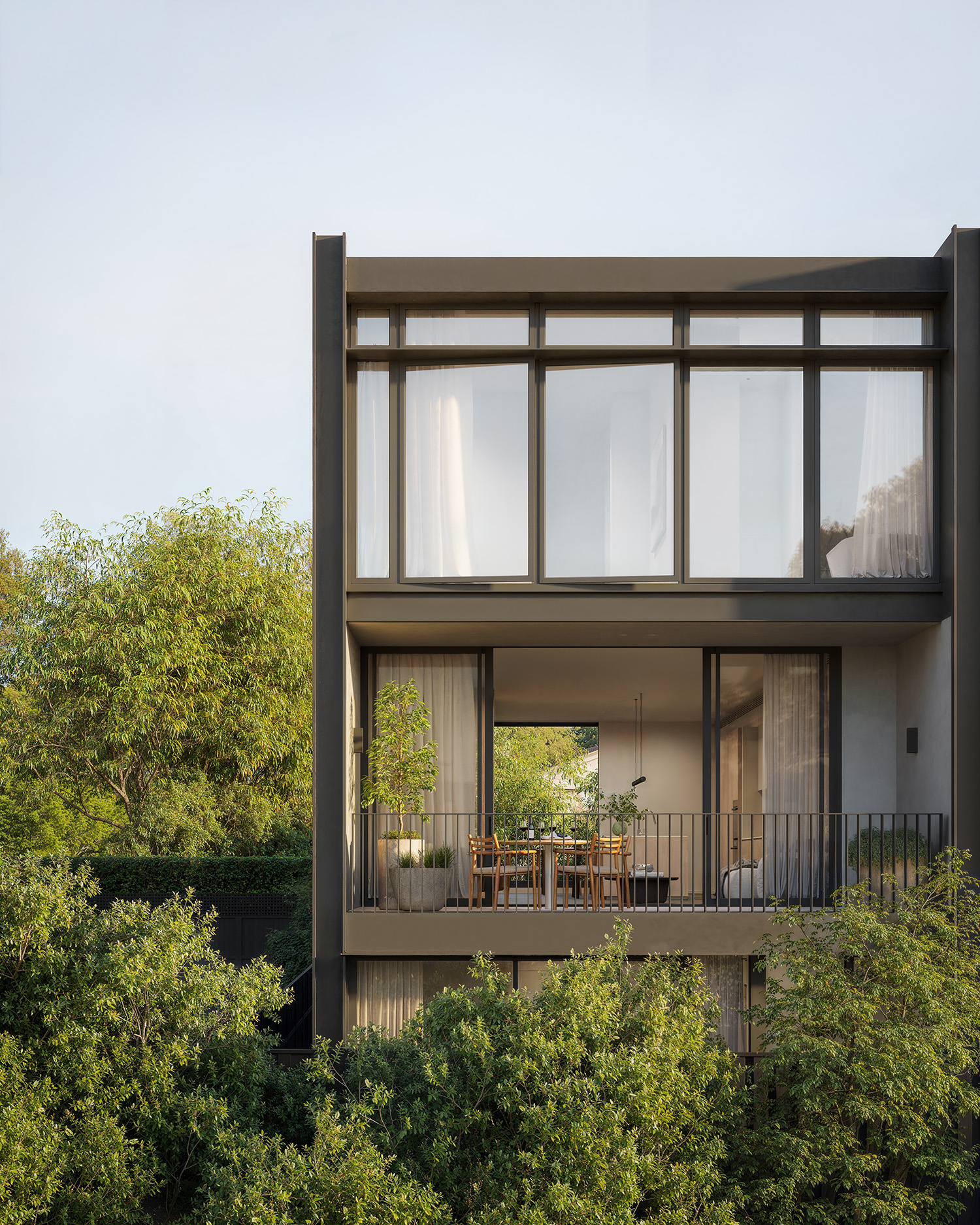 Modern home's upper-level glass facade with an open balcony featuring a small outdoor dining set and potted plants, framed by lush green trees.
