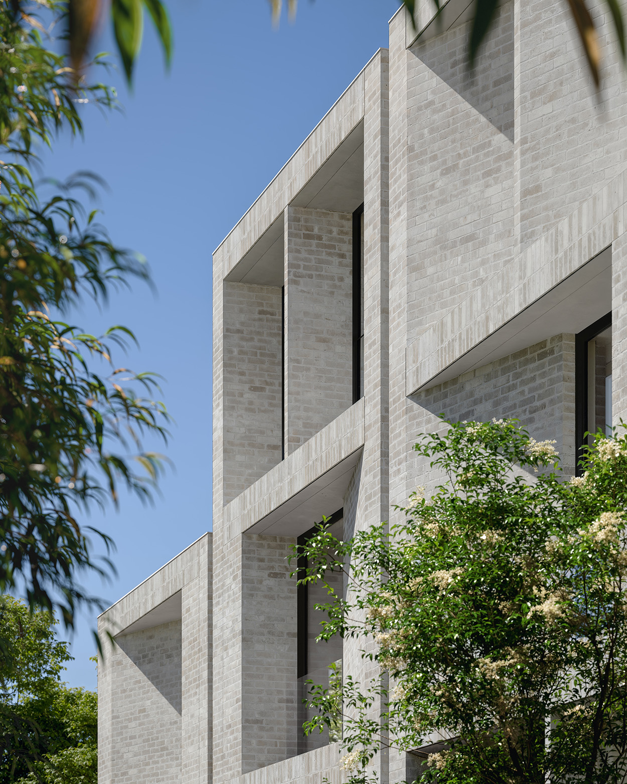 Minimalist pale-brick facade with deep recessed rectangular openings casting sharp geometric shadows, framed by leafy trees and a clear blue sky.