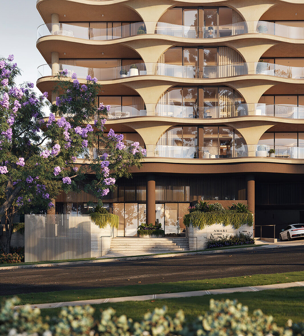 Curved, wave-like concrete balconies form a rhythmic, sunlit facade on a modern residential building, with glass railings, a planted street-level entrance and a purple-flowering tree in the foreground.