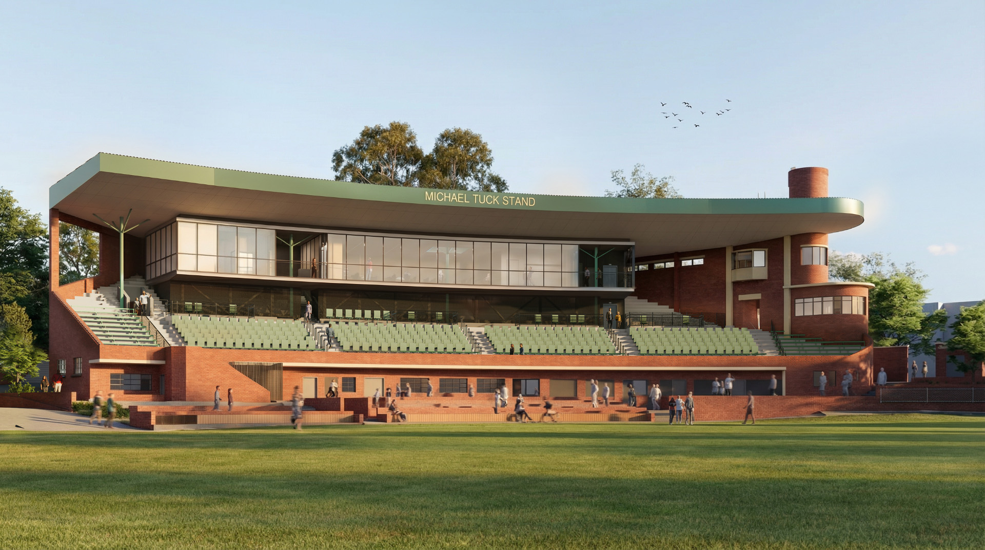 Michael Tuck Stand at Glenferrie Oval: a modern red‑brick grandstand with a green cantilevered roof, tiered green seating, glassed VIP boxes, people on terraces and concourses, and a wide grassy playing field in the foreground.